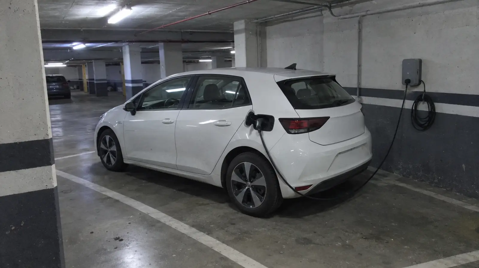 Coche eléctrico blanco cargando en su plaza de un garaje subterráneo, conectado a un cargador gris fijado a la pared