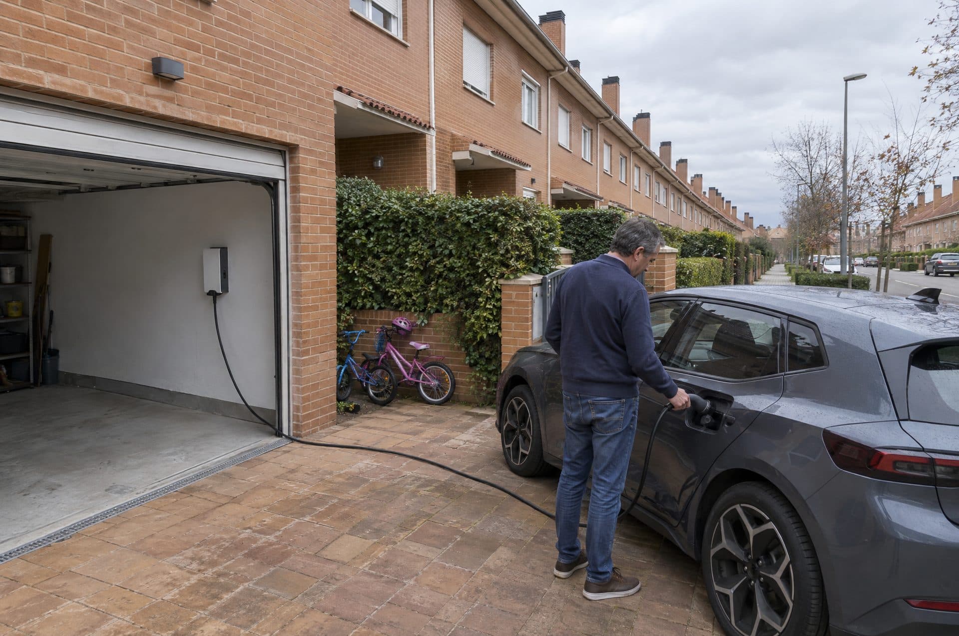 Vecino enchufando su coche eléctrico frente a un adosado de ladrillo con el cargador dentro del garaje abierto