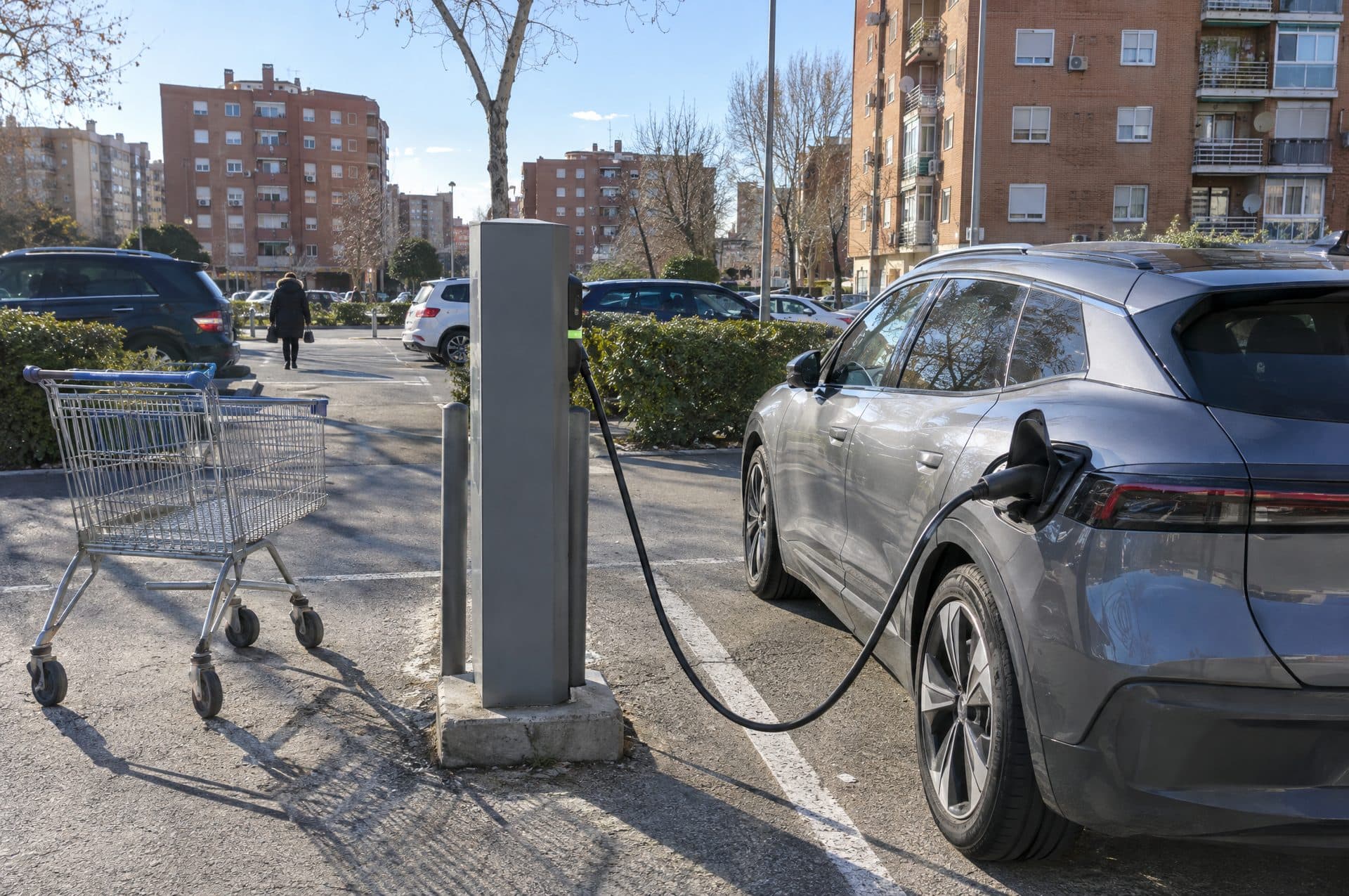 Coche eléctrico cargando en un poste de un aparcamiento de barrio rodeado de bloques de ladrillo, con un carro de la compra al lado