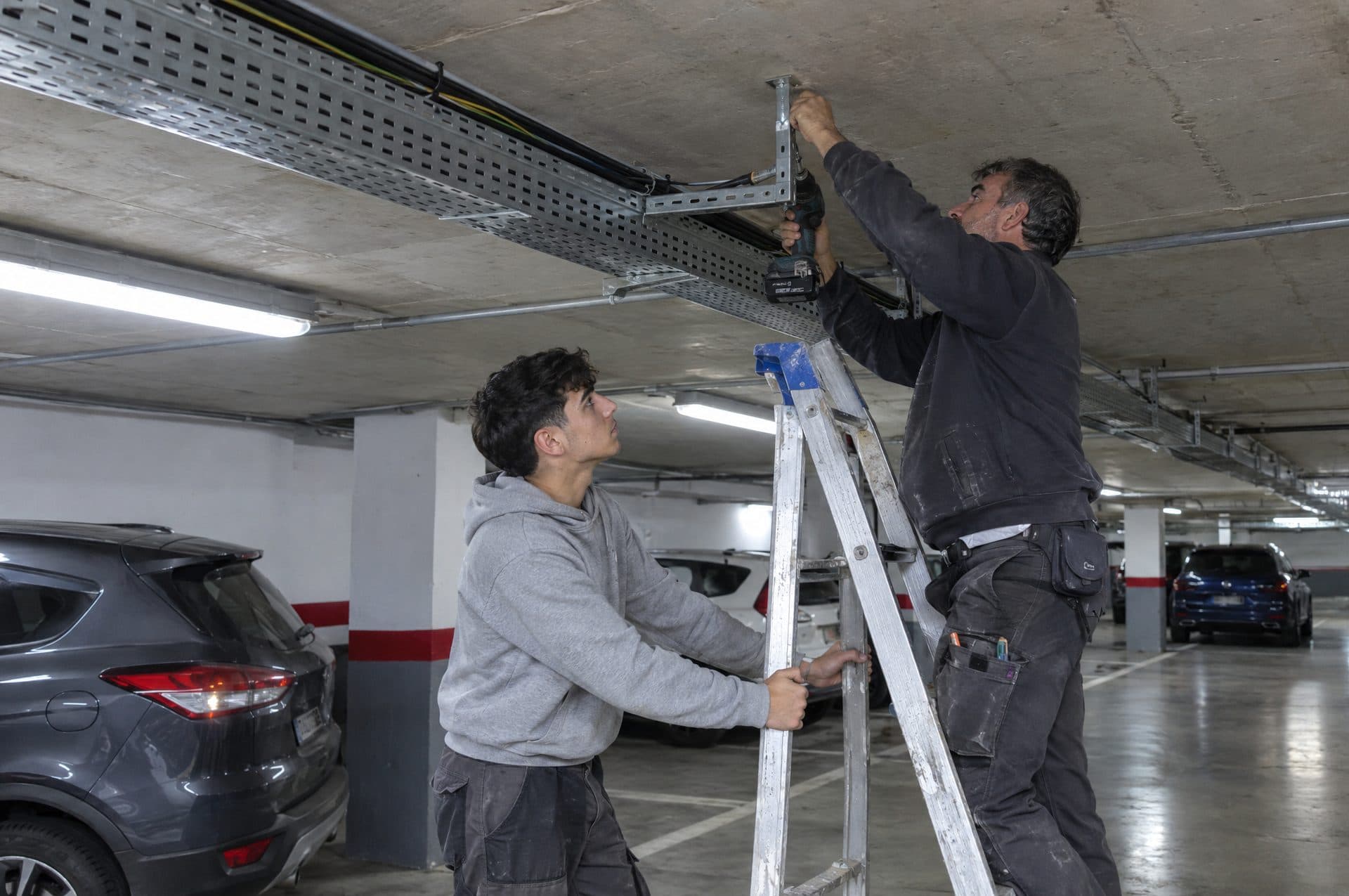 Aprendiz sujetando la escalera mientras un electricista fija un soporte de bandeja al techo de un garaje