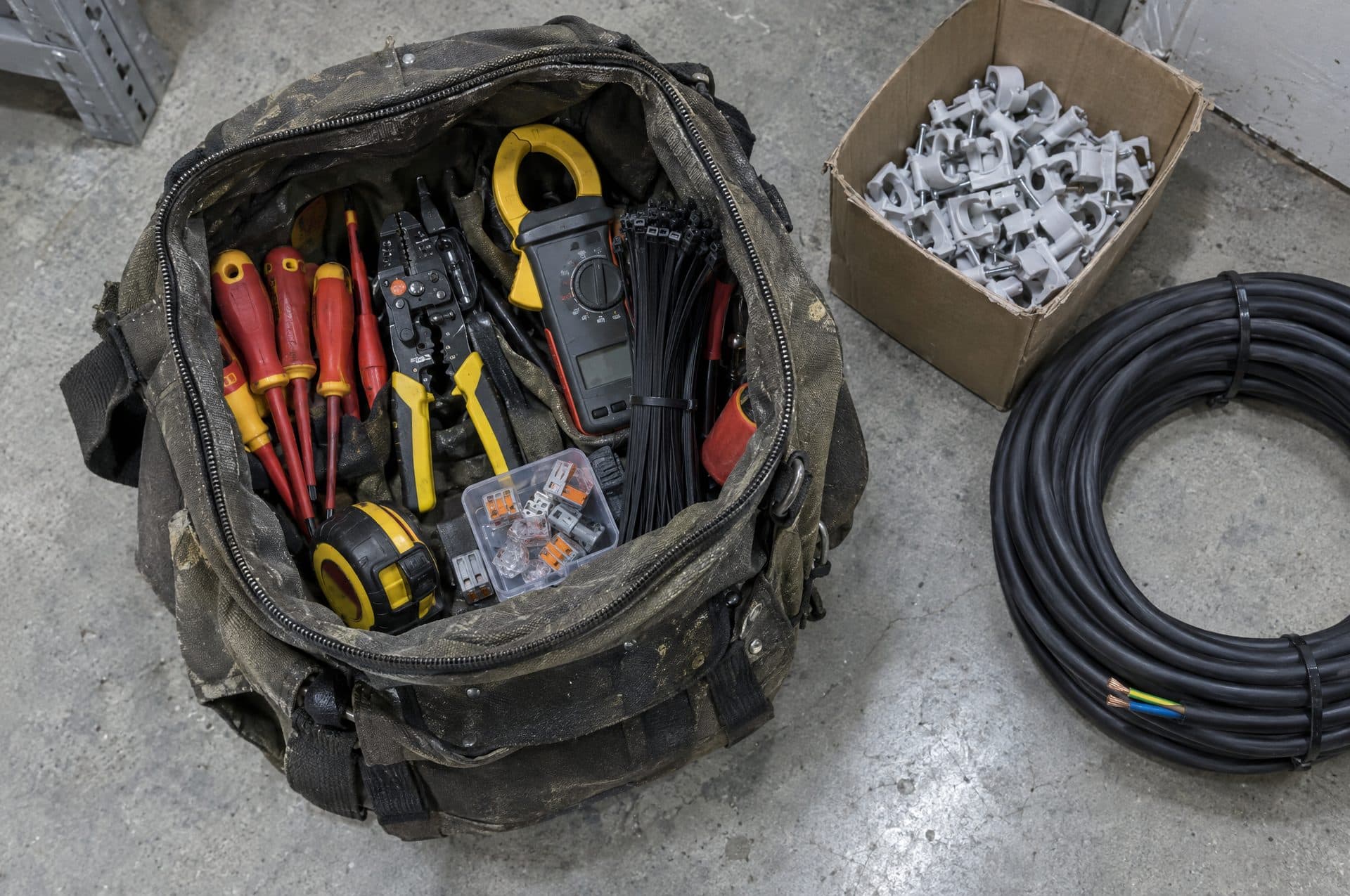 Bolsa de herramientas de electricista con destornilladores aislados, pinza amperimétrica y bridas, junto a grapas y una bobina de cable