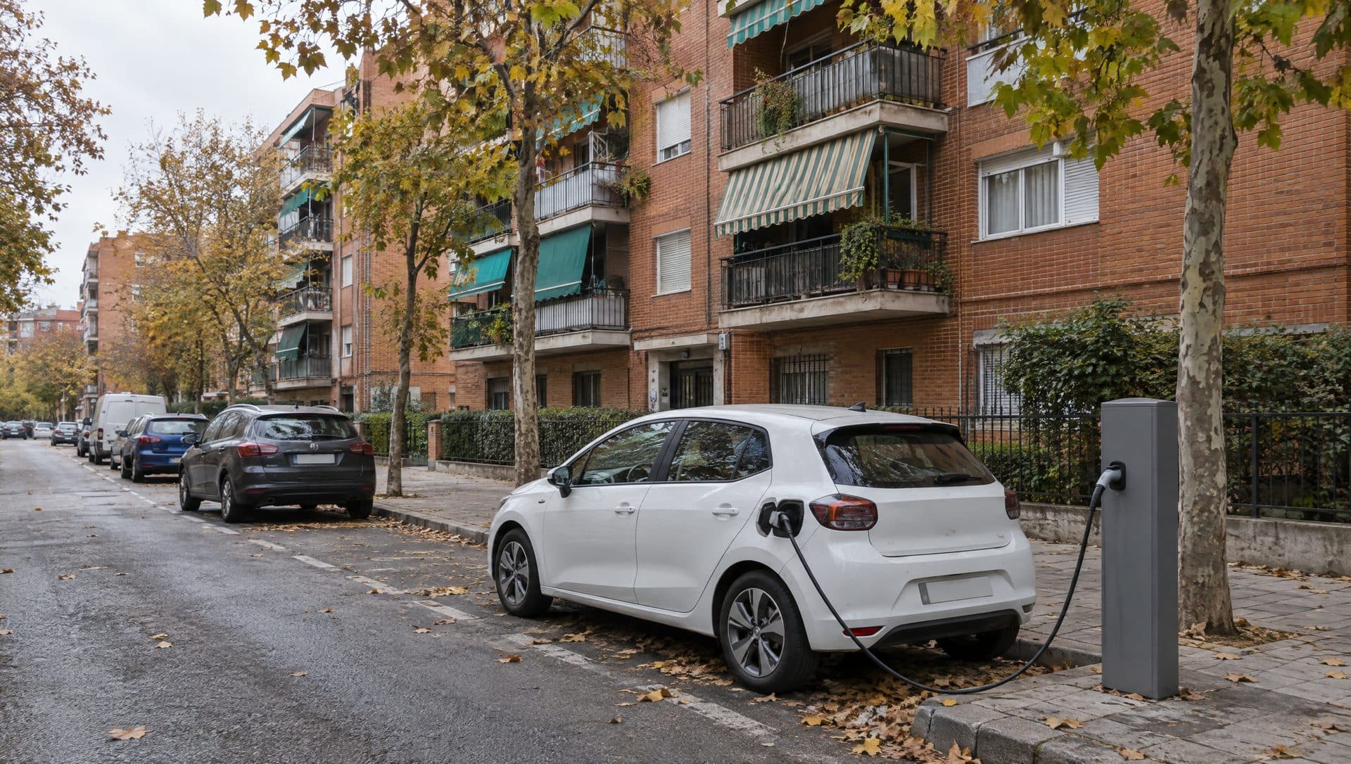 Coche eléctrico blanco cargando en un poste de recarga de calle, frente a bloques de ladrillo con toldos y árboles en otoño