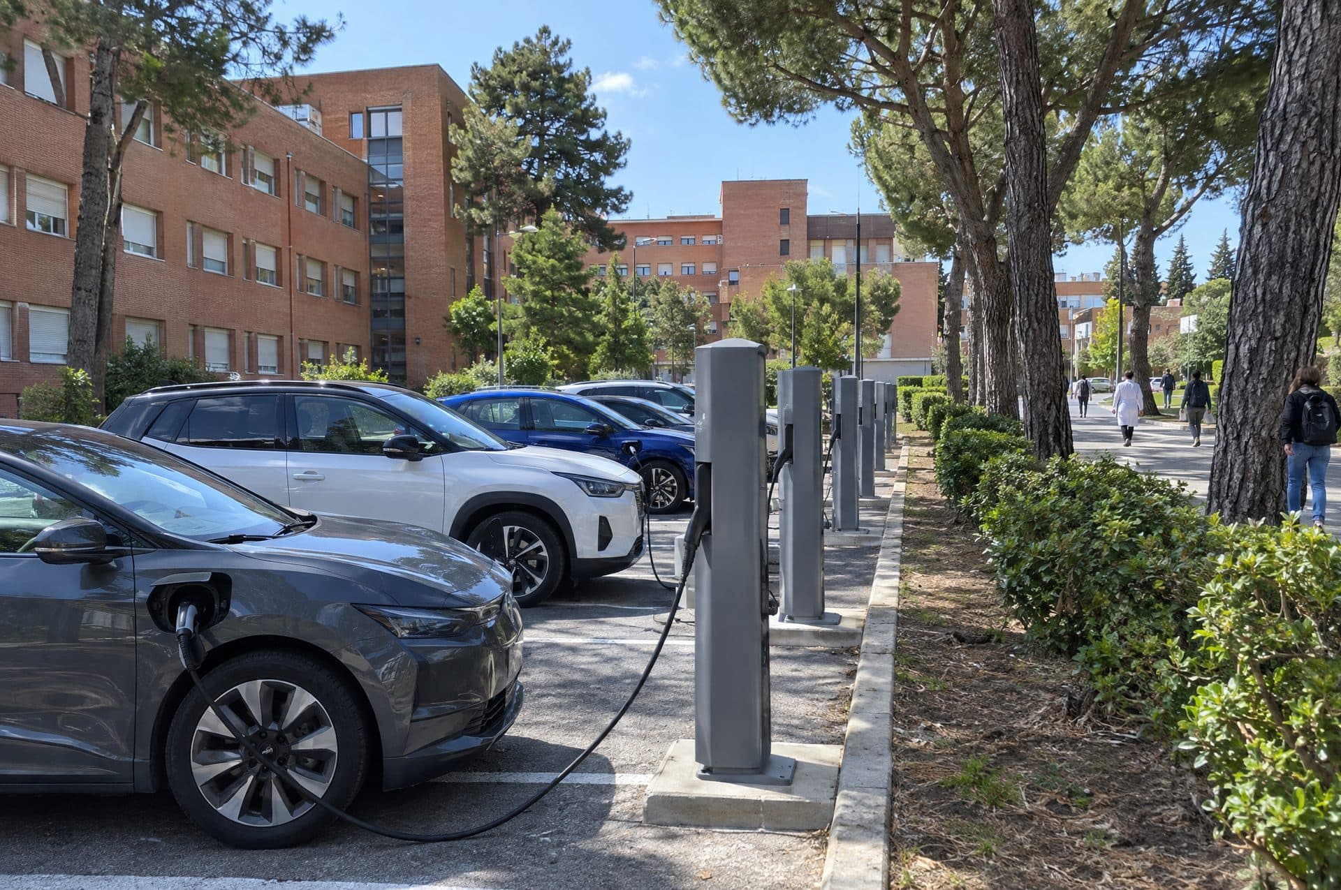Aparcamiento de personal con una fila de postes de recarga y coches eléctricos cargando entre pinos y edificios de ladrillo