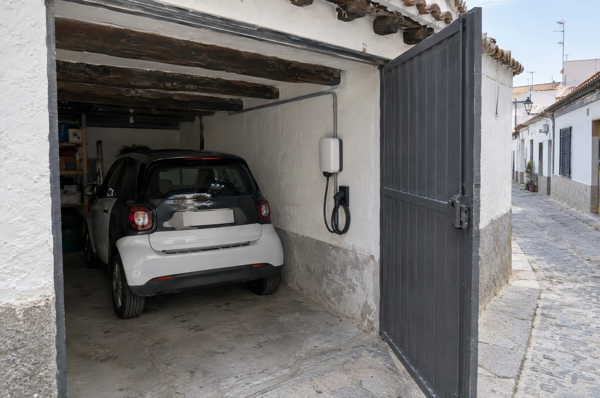 Cochera encalada de una casa baja con vigas de madera, un cargador blanco en la pared y un coche eléctrico pequeño dentro