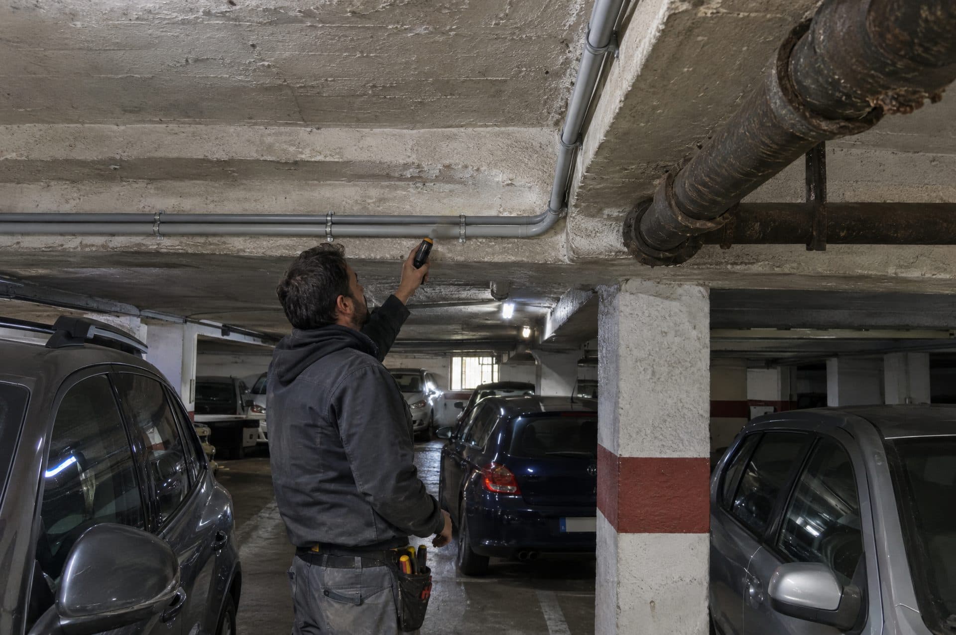 Electricista revisando el recorrido de un tubo gris por el techo bajo de un garaje antiguo con vigas de hormigón