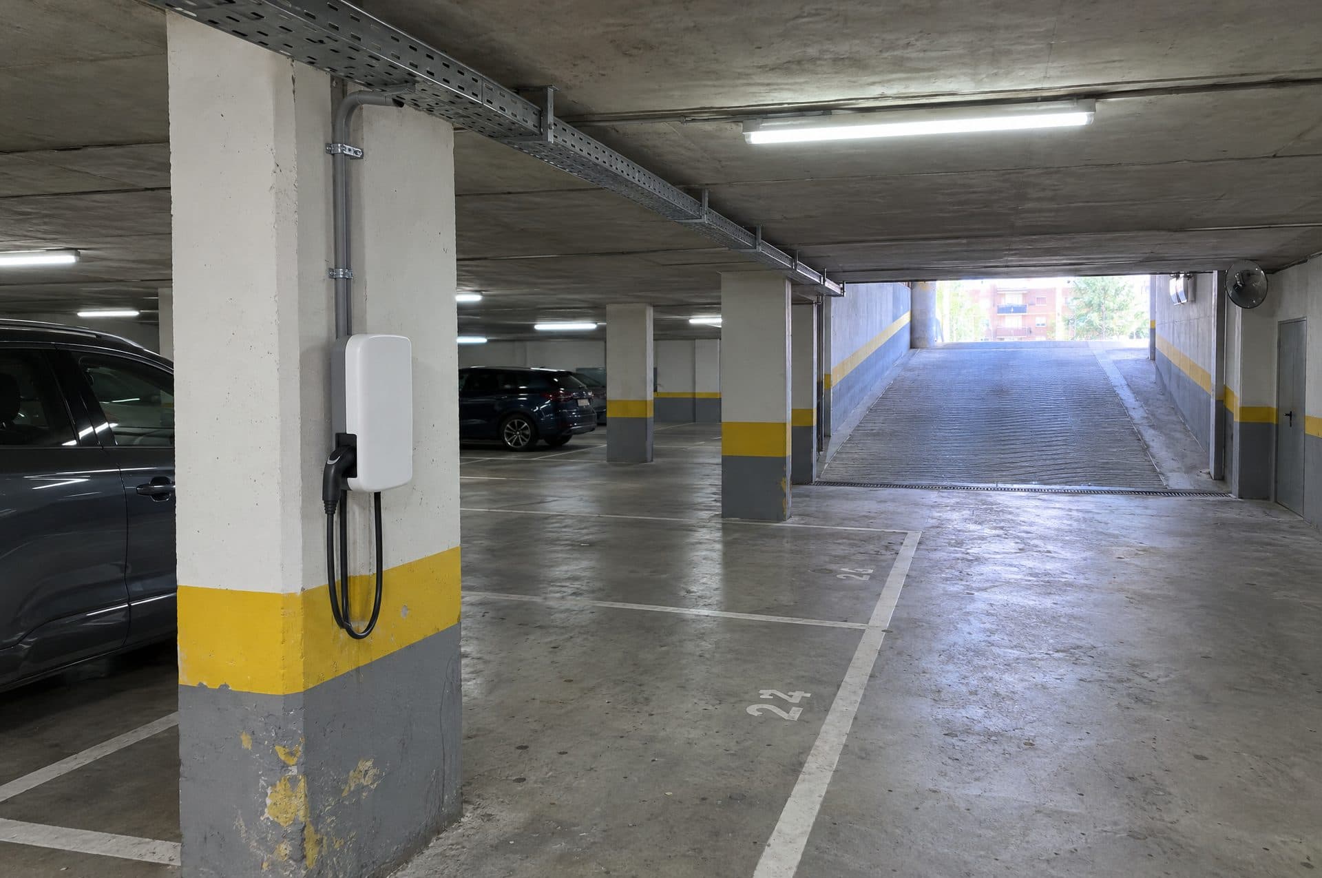Garaje comunitario subterráneo con un cargador blanco en un pilar de franja amarilla, tubo gris hasta la bandeja del techo y la rampa de salida al fondo