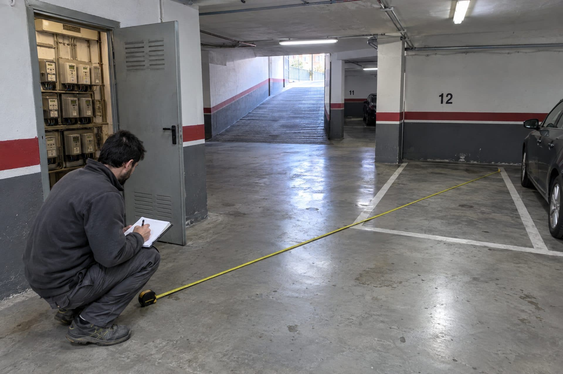 Electricista midiendo con cinta métrica la distancia desde el cuarto de contadores hasta una plaza de garaje y tomando notas