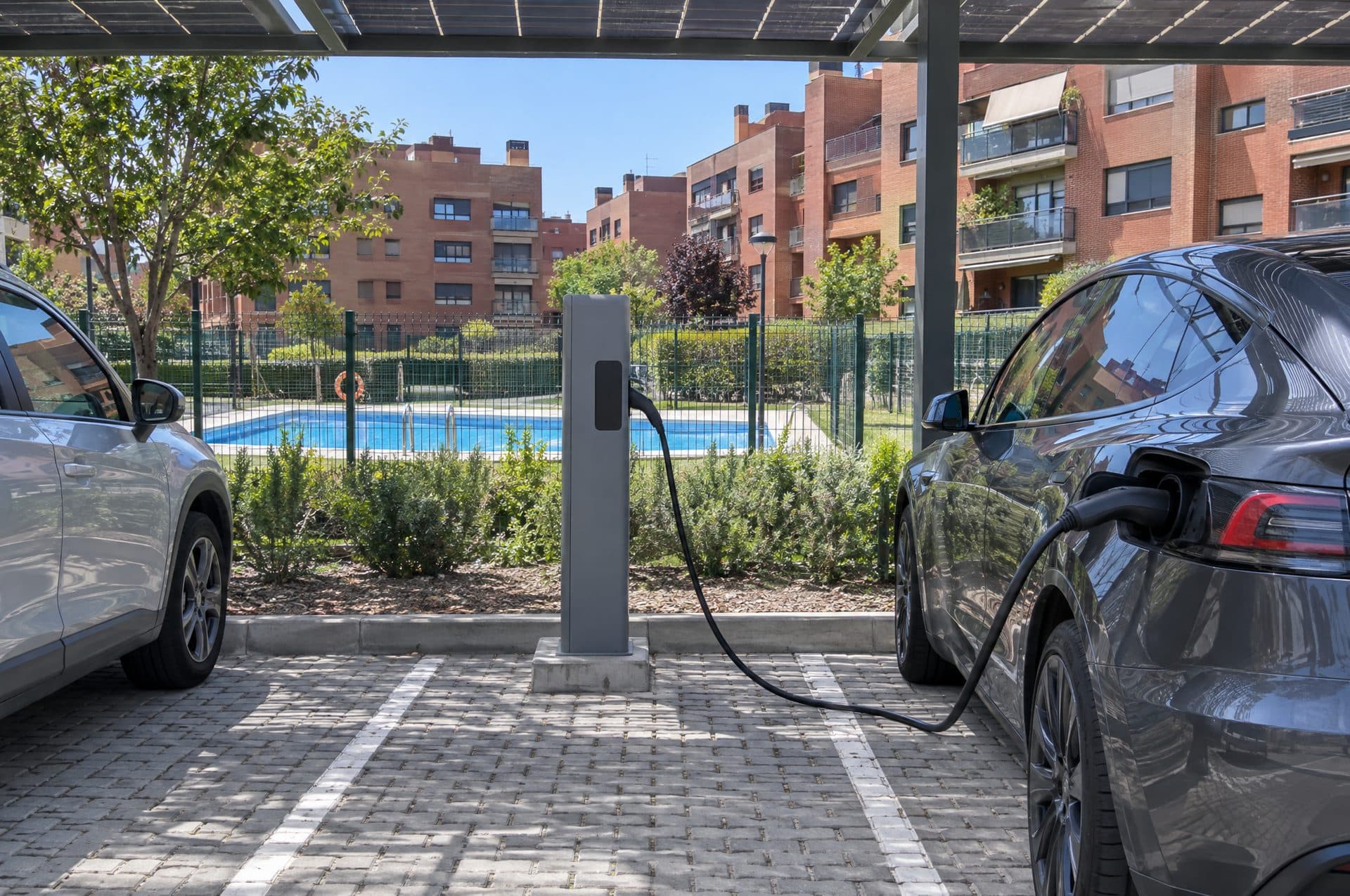 Plaza exterior de una urbanización con un poste de recarga bajo una pérgola, coche cargando y la piscina al fondo