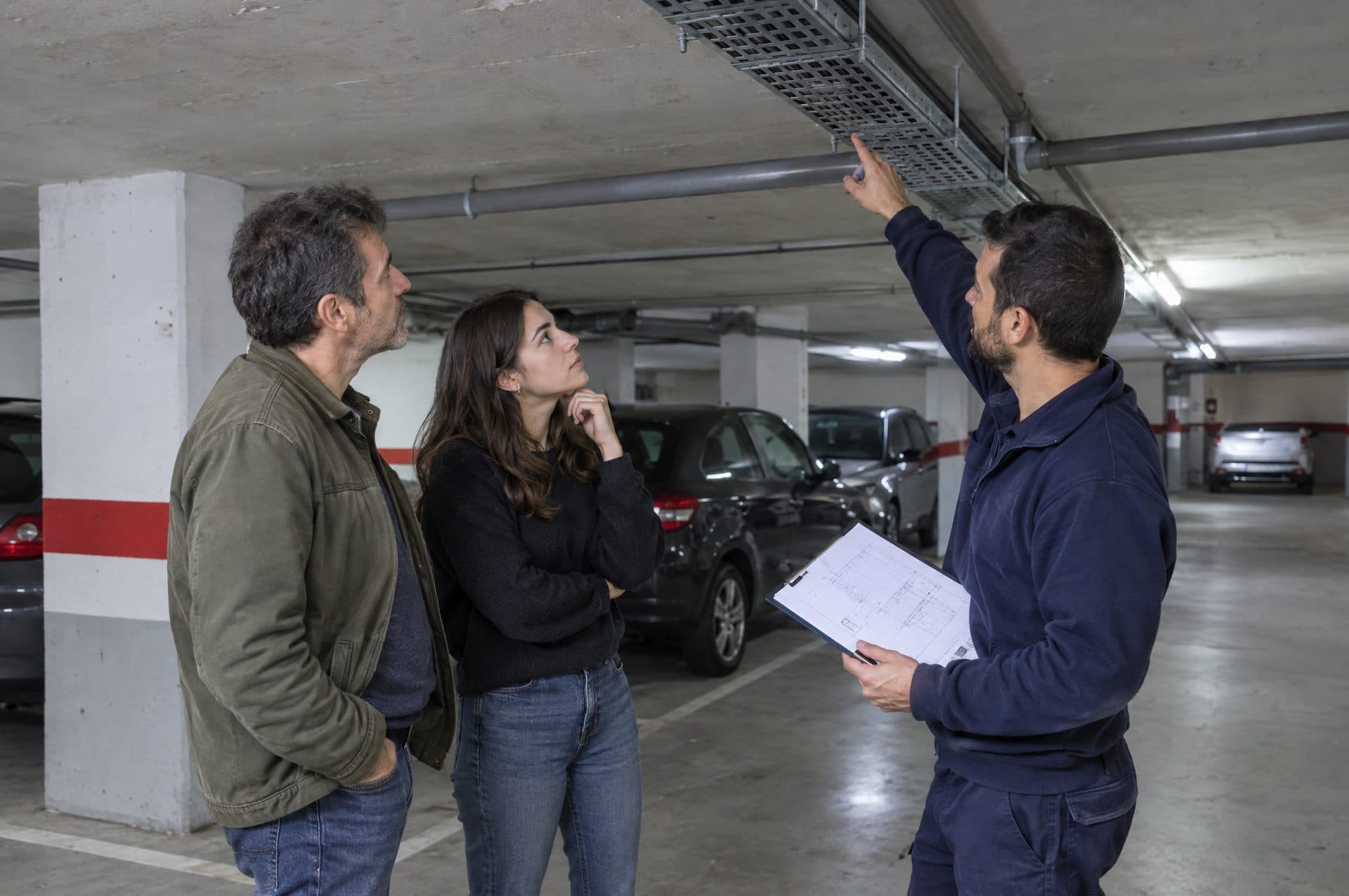 Electricista con una carpeta señalando el techo de un garaje comunitario a dos vecinos para explicar por dónde irá la bandeja