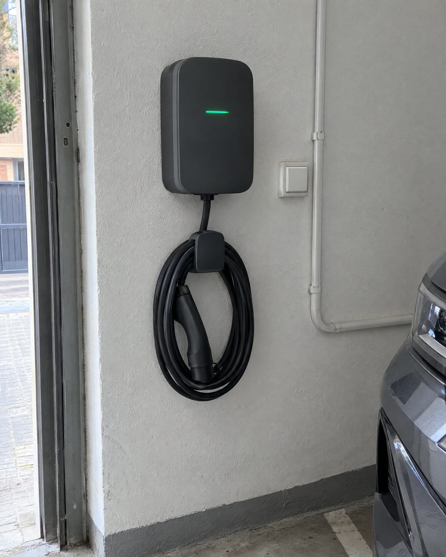 Cargador de pared gris oscuro con el piloto verde encendido y el cable enrollado en su soporte, en la pared de un garaje